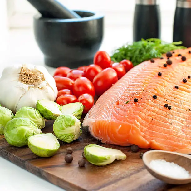 Big piece of fresh raw salmon/trout with vegetables and seasoning on wooden cutting board- ready to eat, ready to cook. Indoors square close-up image