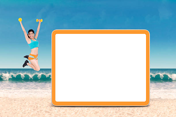 Happy woman wearing sportswear jumps at beach next to empty board while holding two dumbbells