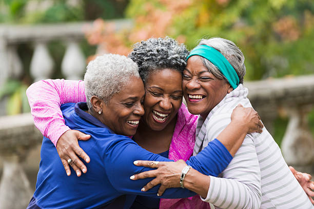 Senior black women hugging A group of three happy senior African American women embracing.  They are standing in a park, arms around each other, smiling and laughing with their eyes closed. cheek to cheek photos stock pictures, royalty-free photos & images