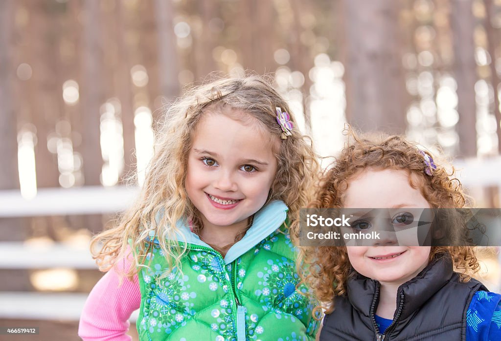 Close-Up of Two Young Sisters Smiling At Camera Close-up of two young girls (sisters) smiling at the camera on a spring day. White rail fence and tall pine trees are visible behind them. Pine Tree Stock Photo