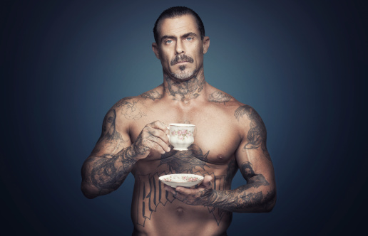 Bare chested adult caucasian man with religious tattoos holding scup of tea. Blue background. Vintage tea cup.