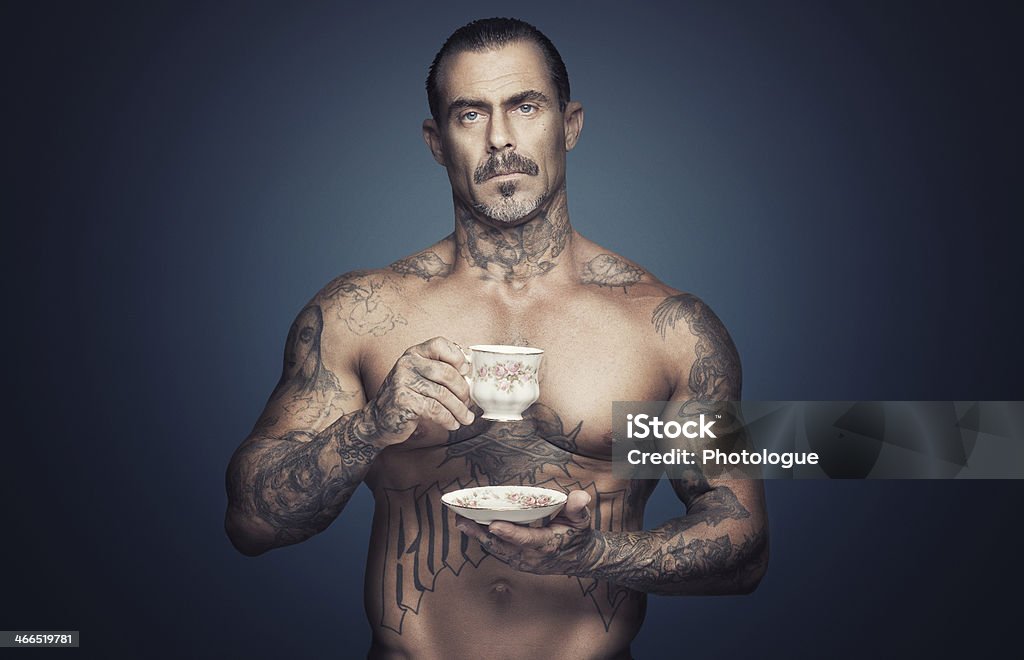 Bare chested man with tattoos holding a cup of tea. Bare chested adult caucasian man with religious tattoos holding scup of tea. Blue background. Vintage tea cup. Men Stock Photo