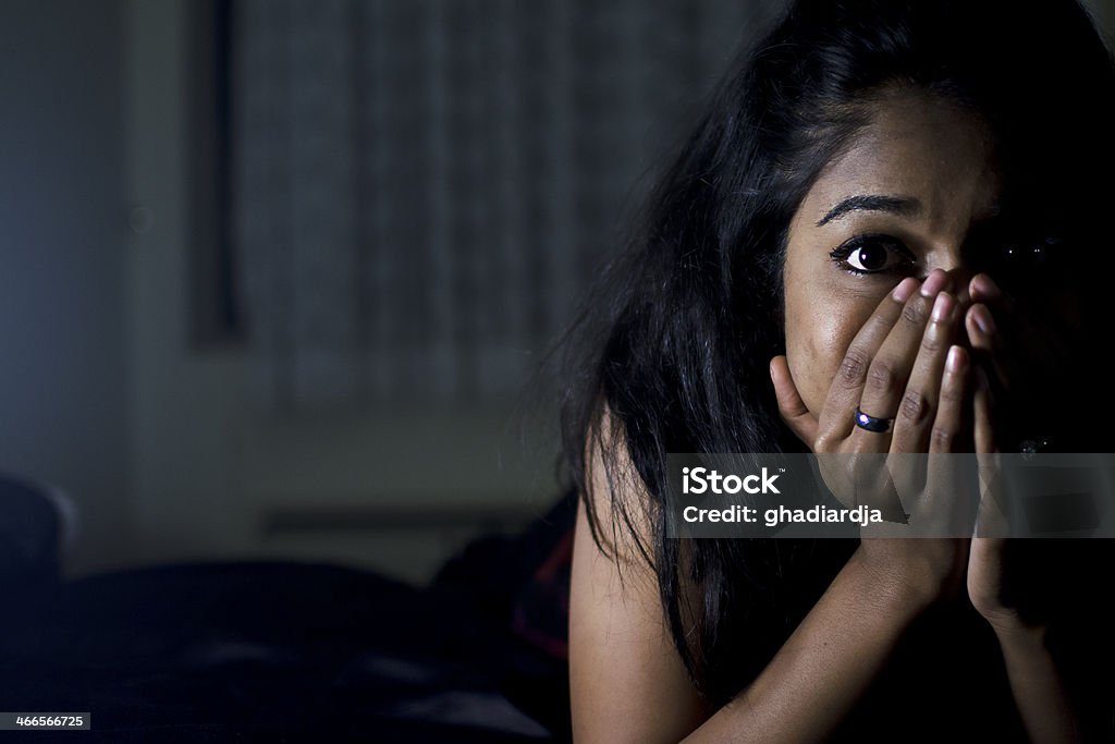 Anxiety A woman lies in a dark bedroom with her mouth covered and looking anxiously into the camera Black Color Stock Photo