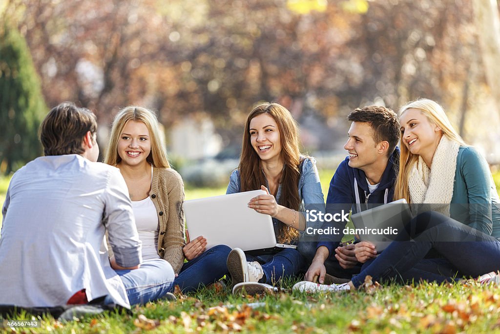 Group of college students studying at campus A group of cheerful university students are having a discussion in a university campus. A group consists of two boys and three girls. A girl with a long red hair in a blue shirt is holding a white laptop. Another girl with long blond hair is holding a digital tablet.  Autumn Stock Photo