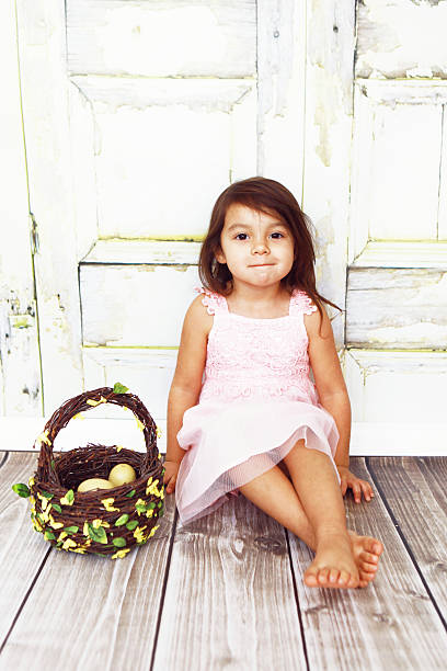 A beautiful little, three years old, girl sits beside her Easter basket. Rustic wooden wall in background. Soft focus. Copy space.