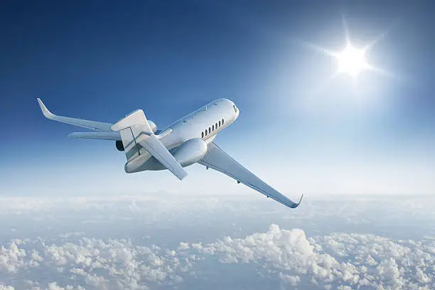 Private jet flies towards the sun in blue sky above the clouds