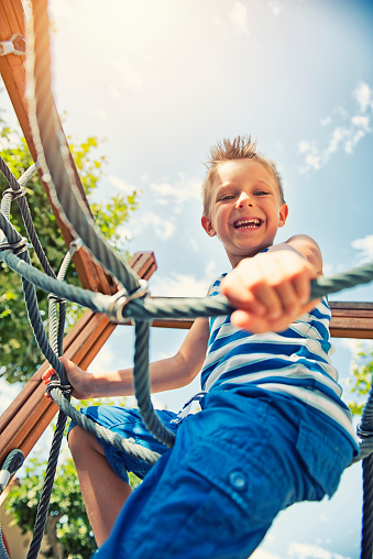 Portrait of little boy climbing in the playground. The boy aged 5 is smiling into the camera. Sunny summer day. Shot from directly below the boy.