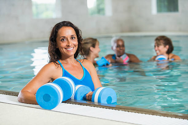 Multi-ethnic group of women in a water aerobics class, in a swimming pool.  They are holding foam dumbbells.  The focus is on the mid adult woman (30s) in the foreground, smiling, looking at camera.  She looks like she might be the teacher or instructor.