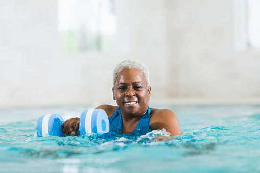 A senior African American woman, in her sixties, doing water aerobics in a swimming pool to stay in shape.  She is holding foam dumbbells in her hands, and smiling at the camera as she does her exercises.  Her head and shoulders are above the water.  She has short, white hair and is wearing a blue swimsuit.
