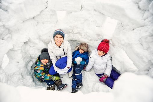Mother and three kids having fun, playing in a snow castle. Girl aged 9 and boys aged 5.