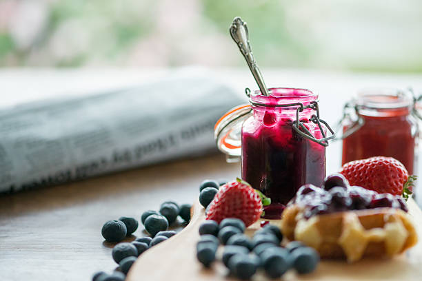 Blueberry, Strawberry Sauces, Waffle, Fresh Fruit, Newspaper, Against Garden Background Breakfast table - Belgium Liege waffles with homemade strawberry and blueberry compote, sauce or jam spooned from opened mason jars.  Fresh blueberries and strawberries on the side.  All set on a wood cutting board.  Drips, spills and a little messy.  Closeup with newspaper and reading glasses in the midground and a flower garden in the background. compote stock pictures, royalty-free photos & images