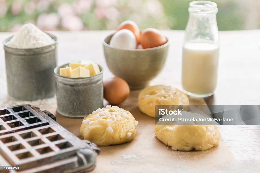 Belgium Waffle Iron and Ingredients, Flour, Butter, Eggs, Milk, Dough Worn waffle iron and Belgium Liege waffle ingredients including, flour, milk, eggs, butter and dough with pearl sugar on parchment paper.  Pearl sugar is used for making authentic Belgium Liege waffles.  Some spills and a little messy.  Close up and view of flower garden in the background.  Country style. American Culture Stock Photo