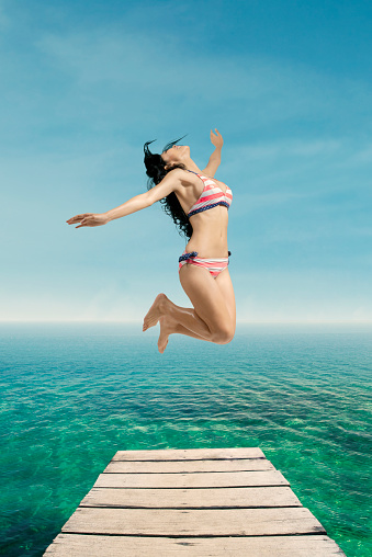 Portrait of joyful woman enjoy freedom at beach by jumping on the pier while wearing bikini