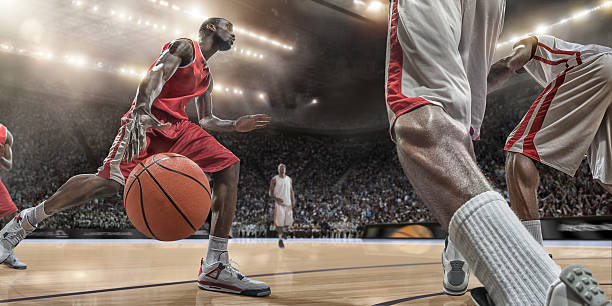 Basketball Player in Action A close up image from a low angle viewpoint of a basketball player dribbling basketball in front of rival players, in mid action. The action takes place in generic indoor floodlit basketball arena full of spectators. All players are wearing generic unbranded basketball kit, and all stadium advertising is fake.  dribbling stock pictures, royalty-free photos & images