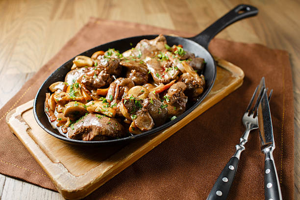 Liver baked with mushrooms, bacon and herbs Liver baked with mushrooms, bacon and herbs in a frying pan on a wooden board closeup close lie knife and fork animal liver stock pictures, royalty-free photos & images