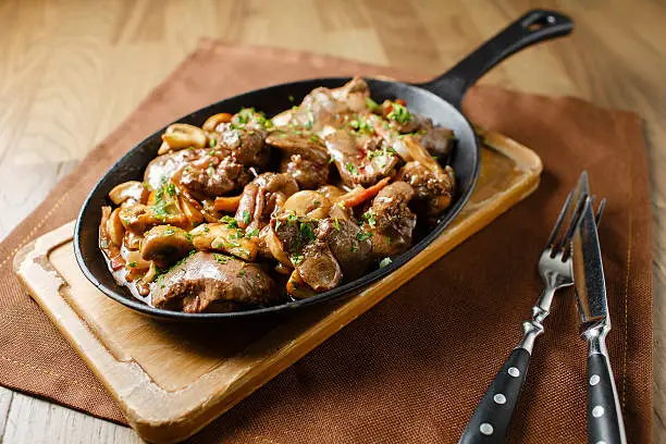 Liver baked with mushrooms, bacon and herbs in a frying pan on a wooden board closeup close lie knife and fork