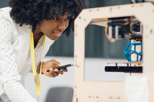 Young smiling african woman with cute curly hair holding her smart phone and looking at 3d printer printing her new project. Selective focus to her positive expression.