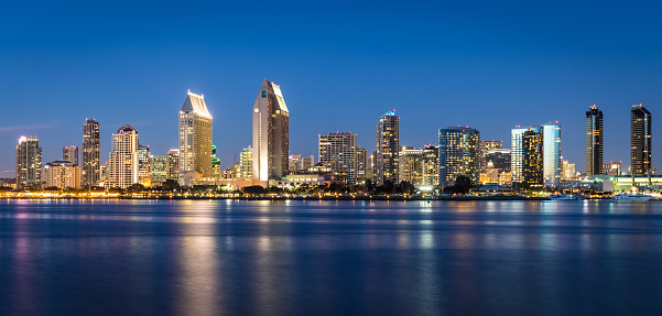 City of San Diego from across San Diego Bay at Coronado island at dusk.  A clear sky without any cloud on a warm summer day. The buildings are lightened and some are reflecting on the water. More than 3 logo visible.
