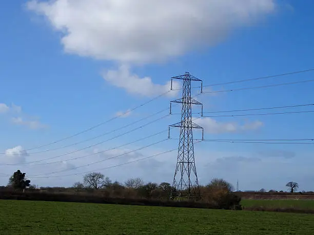 Photo showing a tall high-voltage transmission tower, which is known in the UK and much of Europe as an electricity pylon.  The photo shows a tower in a countryside field, with conductors, insulator strings and cables / wires stretching into the distance.  This part of the structure is close to the top (peak) and known as the 'cage'.
