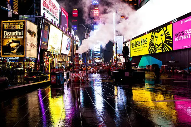 View of Times Square at night illuminated with neon colors. Wet streets. Broadway plays and chain store ads on billboard. Smoke rises from ground. New York city.