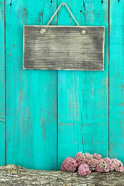 Blank rustic sign hanging on antique teal blue wood fence with log and flower border