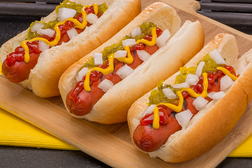 Three Hot Dogs with Mustard, Ketchup, pickle relish and onions on a wood cutting board 