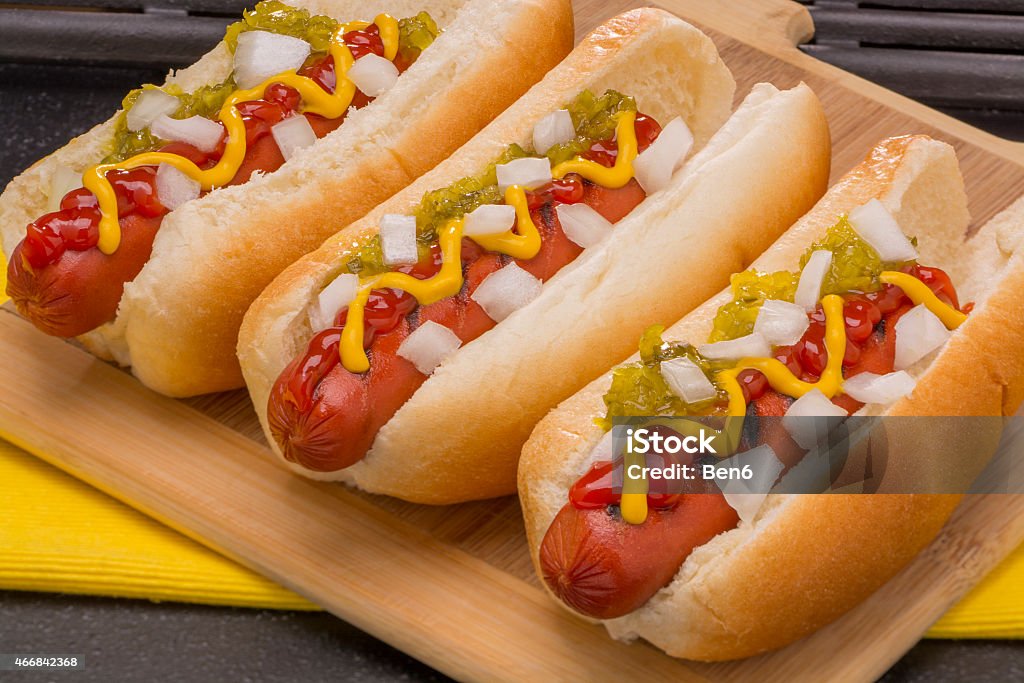 Hot Dogs Ready to Eat Three Hot Dogs with Mustard, Ketchup, pickle relish and onions on a wood cutting board  Hot Dog Stock Photo