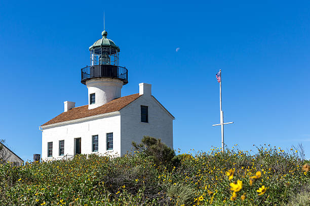 The Historic Lighthouse in Point Loma, San Diego, California. It's a beautiful day of summer with a clear sky, the moon is still visible and there is a us flag floating with the wind and yellow flowers in the foreground.