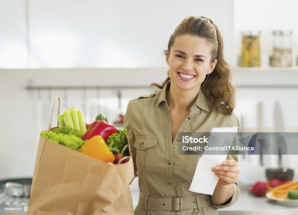 Smiling housewife with check and shopping bag full of vegetables Smiling young housewife with check and shopping bag full of vegetables Adult Stock Photo