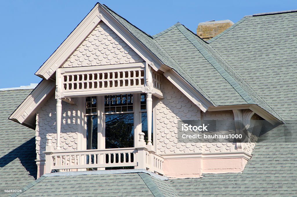 Victorian Architectural Detail with Gingerbread Shakes Architectural detail of a large Victorian-era dormer with columns and gingerbread shake siding extending from the roof line of a large home. Bright light with contrasting shadows creates an interesting pattern with the roof lines. Pink paint with a grey roof against a blue sky. 2015 Stock Photo