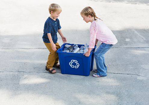 A young girl and her little brother standing on the driveway with a blue recycling bin full of empty bottles and cans.  They are helping to bring the bin out to the curb for collection, about to lift it up to carry.  The boy is 4 years old and his sister is 6, both with blond hair.