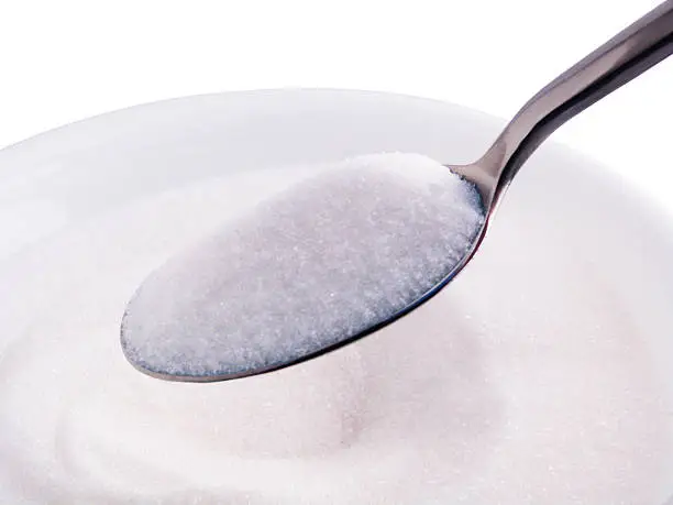 A heaping silver spoonful of white granulated sugar suspended over a white bowl of sugar, isolated on a white background.