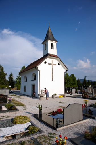 Bled, Slovenia - July 4, 2012: Picturesque church in Slovenian countryside at evening. A senior woman is filling water into a flowerpot