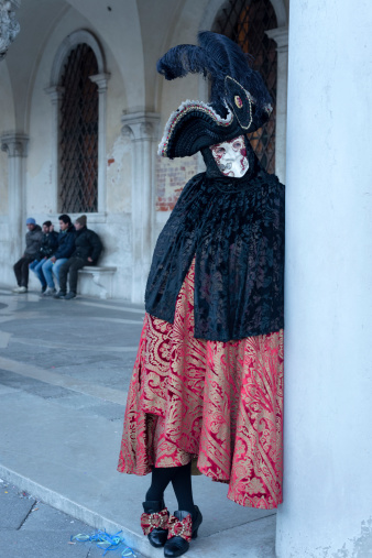 Venice, Veneto, Italy - February 8th, 2013: Black and red mask, participant of the 2013 Carnival celebrations, posing for casual photographers and tourists on St. Mark's Square, by the Doges Palace. Four men sitting by the big window in the background.