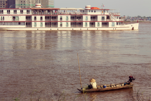 Phnom Penh, Cambodia - December 4, 2013: A man fishing from a small boat by the riverside watches a river cruise ship with tourists pass by.