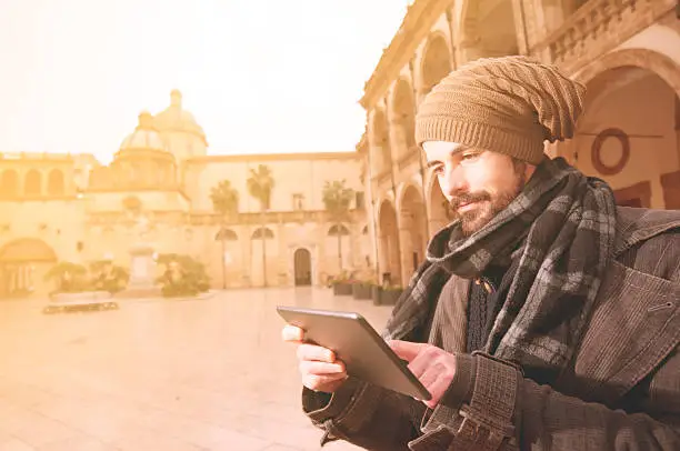 Young Man reading a tablet in a euroepan square with a church on the background in instagram toned style