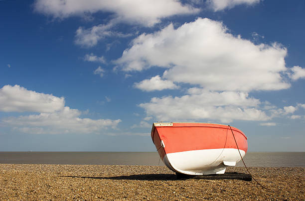 A red and white boat on the shingle at Dunwich, on the Suffolk Heritage Coast, in the Suffolk Coast and Heaths Area of Outstanding Natural Beauty. On this bright, sunny day at the end of February, fluffy white clouds and a hint of moon feature in the blue sky above calm seas. Polarizing filter used.