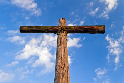An old rugged wooden cross stands against a blue sky draped with white clouds as viewed from below and in front