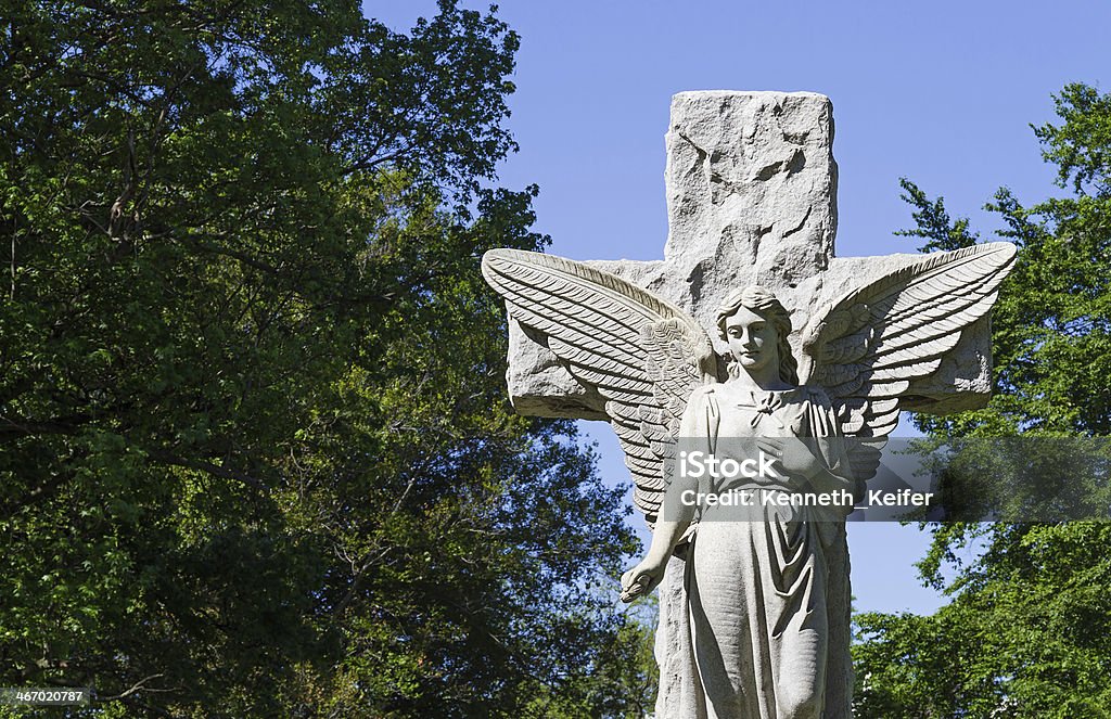 Cemetery Angel A stone angel and cross statue, typically found in many graveyards and cemeteries  stands with green trees and a blue sky behind. Angel Stock Photo