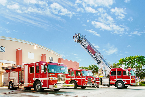 Three fire engine emergency vehicles stand in front of the fire house with the ladder of the hook and ladder truck in an up position for inspection.