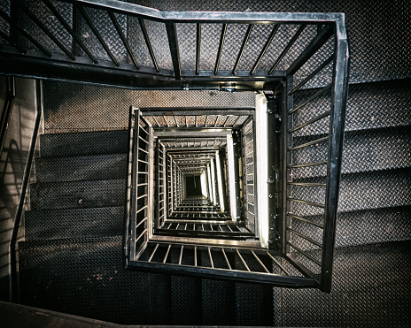A view from the top, looking down into an industrial building stairwell. Metal stairs and railings lead down several floors into the basement. 