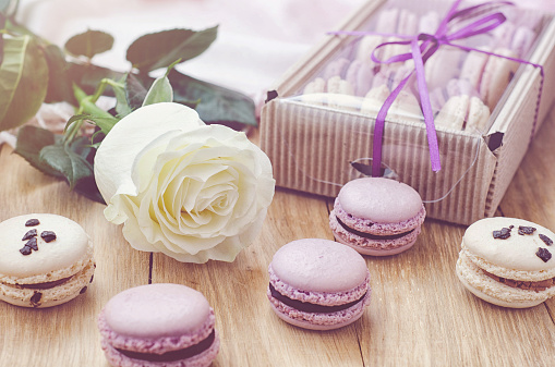 Soft and tasty macaroons lilac and beige colors with a white rose and gift box for valentine's day or mother's day. Everything lying on a wooden surface. Photo toned in light hazy lilac color. Selective focus