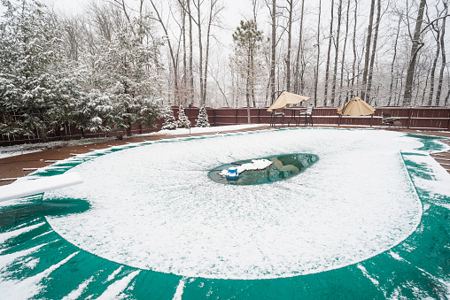 Inground swimming pool with cover on it and water pump during a snowfall in winter, Indiana, USA