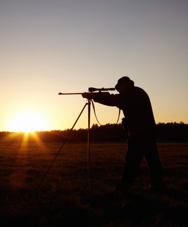 A silhouette of a man aiming with his rifle that is held up by a tripod in the outdoors at sunrise