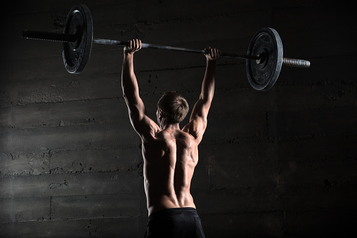 Portrait of a handsome athlete from behind. Athlete raises the barbell over your head. Studio shots in the dark tone.