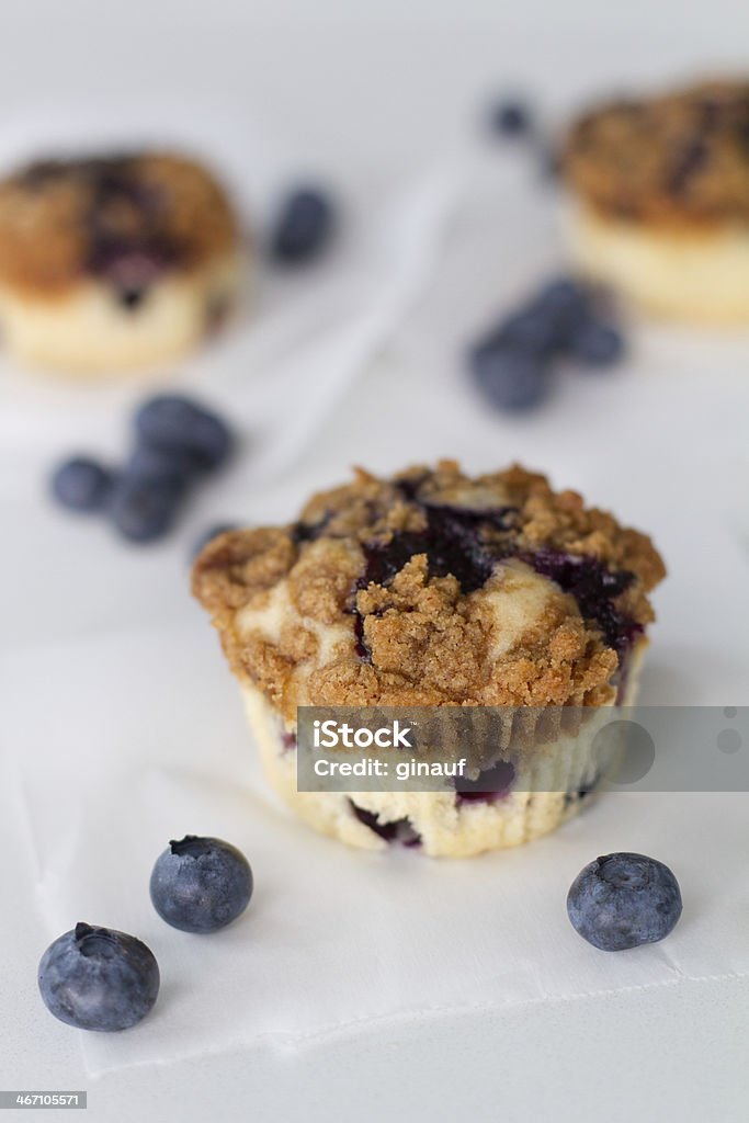 blueberry muffins A photograph of blueberry muffins with a streusel topping, placed on parchment paper on a white countertop.  Fresh blueberries are scattered throughout the counter's surface. Baked Stock Photo