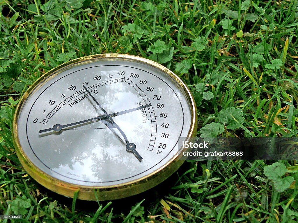 Thermometer In Grass A thermometer and barometer lies outdoor in a green meadow. Some trees are reflecting in the glass surface of the round and flat technical equipment with two indicators pointing into different directions. 2015 Stock Photo