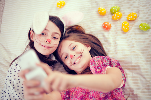 Cute girls friends having for for Easter time .Wearing rabbit ears, lying on floor with coloured Easter eggs around them taking selfie