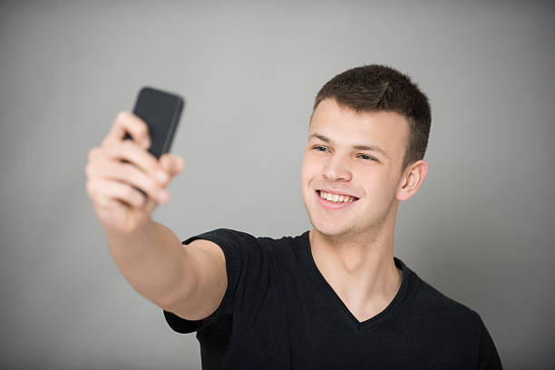 Happy teenage boy in V-neck black t-shirt taking self portrait with smart phone over gray background