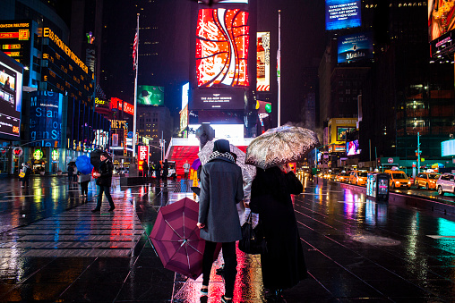 New York City, NY, USA - March 10, 2015: Women tourists take in the scene in Times Square. Steady rain falls. Army of yellow cabs  flows down 7th ave. Electronic billboards and jumbotrons advertise Coca Cola, Budweiser, Morgan Stanley, m&m's, Oreo cookies and more. Some carry umbrellas, others go without.